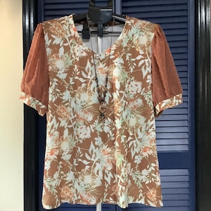 May include: A short-sleeved blouse featuring a brown base with a white and light green floral pattern, complemented by sheer brown puff sleeves with a subtle dotted texture and a V-neckline, displayed with a long beaded pendant necklace.
