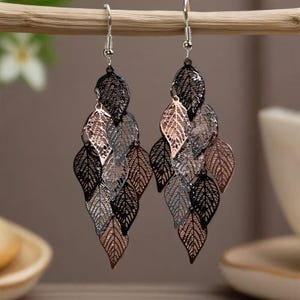 May include: A pair of dangling earrings featuring a leaf design. The earrings are made of metal and have a mix of black, silver, and rose gold tones. The leaves are intricately cut out, creating a delicate and airy look.