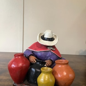 May include: A colorful ceramic figurine of a person wearing a white hat with a black band and a red cape, holding several small, painted vases. The vases are in red, black, yellow, and orange. The figurine is on a dark brown surface.