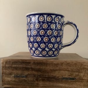May include: A ceramic mug featuring a repeating pattern of blue and white geometric floral shapes with small brown circular centers. The mug has a curved handle on the right side and a smooth white interior rim, showcasing a classic glazed finish.