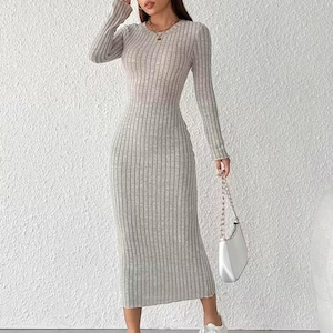 May include: A light gray, ribbed-knit midi dress with long sleeves. The dress has a form-fitting silhouette and a round neckline. The model is wearing white sneakers and carrying a white handbag with a chain strap. The background is a textured white wall.