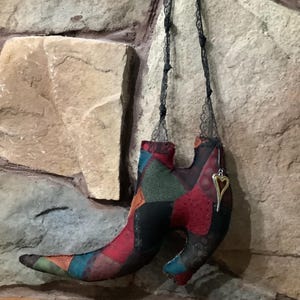 May include: A fabric shoe ornament constructed from various patchwork pieces in shades of red, black, green, and blue. The shoe features a pointed toe and a curved heel, suspended by a thin black lace strap with a silver-toned heart charm attached.