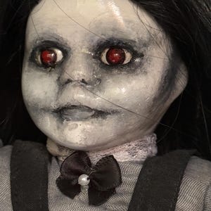 May include: A porcelain doll with a pale face, black hair, and red eyes. The doll is dressed in a grey dress with black detailing and a black bow with a pearl. The doll has a spooky, haunted appearance.