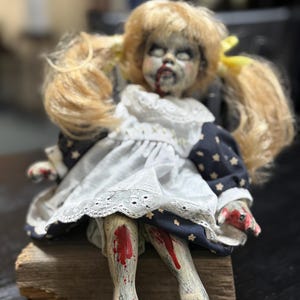 Puede incluir: Una muñeca espeluznante con coletas rubias, vestida con un vestido blanco con un corpiño azul marino y un estampado de estrellas. La muñeca tiene una cara pálida con sangre alrededor de la boca y en las piernas y manos. La muñeca está sentada en un bloque de madera.