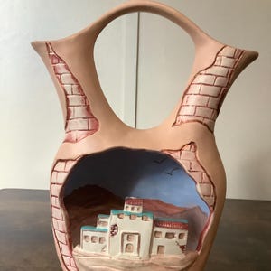 Puede incluir: Un jarrón de cerámica color melocotón con asa, con un diseño de estilo suroeste. El jarrón tiene un recorte que revela un paisaje desértico pintado con edificios. El exterior tiene un patrón similar a ladrillos.