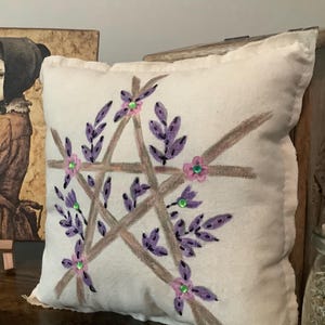 May include: A square, off-white pillow with a hand-painted pentagram design. The pentagram is formed by brown lines and adorned with purple leaves, pink flowers, and green gem accents. The pillow has a soft, textured appearance.
