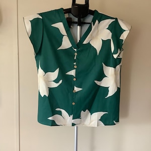 May include: A dark green short-sleeved blouse featuring a large white floral pattern. The garment has a V-neckline and a front button-down closure with five light-colored circular buttons. The sleeves are short and slightly flared at the edges.