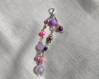 May include: A beaded charm with a silver clasp, featuring a cascade of colorful beads. The beads are in shades of pink, purple, gold, and brown. The charm is likely for a keychain or jewelry, adding a touch of color and personality.