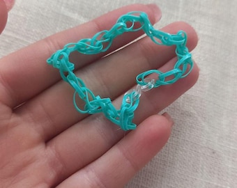 May include: A turquoise rubber band bracelet with a chain link design. The bracelet is made of small, connected rubber bands. The bracelet is held in a hand against a neutral background.