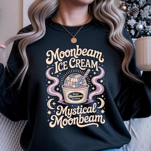 Op de afbeelding: Zwarte sweatshirt met een kleurrijke afbeelding met de tekst "Moonbeam Ice Cream Mystical Moonbeam". Het ontwerp toont een bolletje ijs in een beker met een lepel, een wereldbol en wervelende paarse accenten. De tekst is in een grillig lettertype.