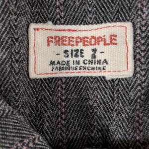 Peut inclure: Gros plan sur une &eacute;tiquette de tissu avec la marque "FREE PEOPLE" en lettres rouges. L'&eacute;tiquette indique &eacute;galement "SIZE 2" et "MADE IN CHINA FABRIQUE EN CHINE" en texte noir. Le tissu pr&eacute;sente un motif &agrave; chevrons gris et noir avec des coutures roses.