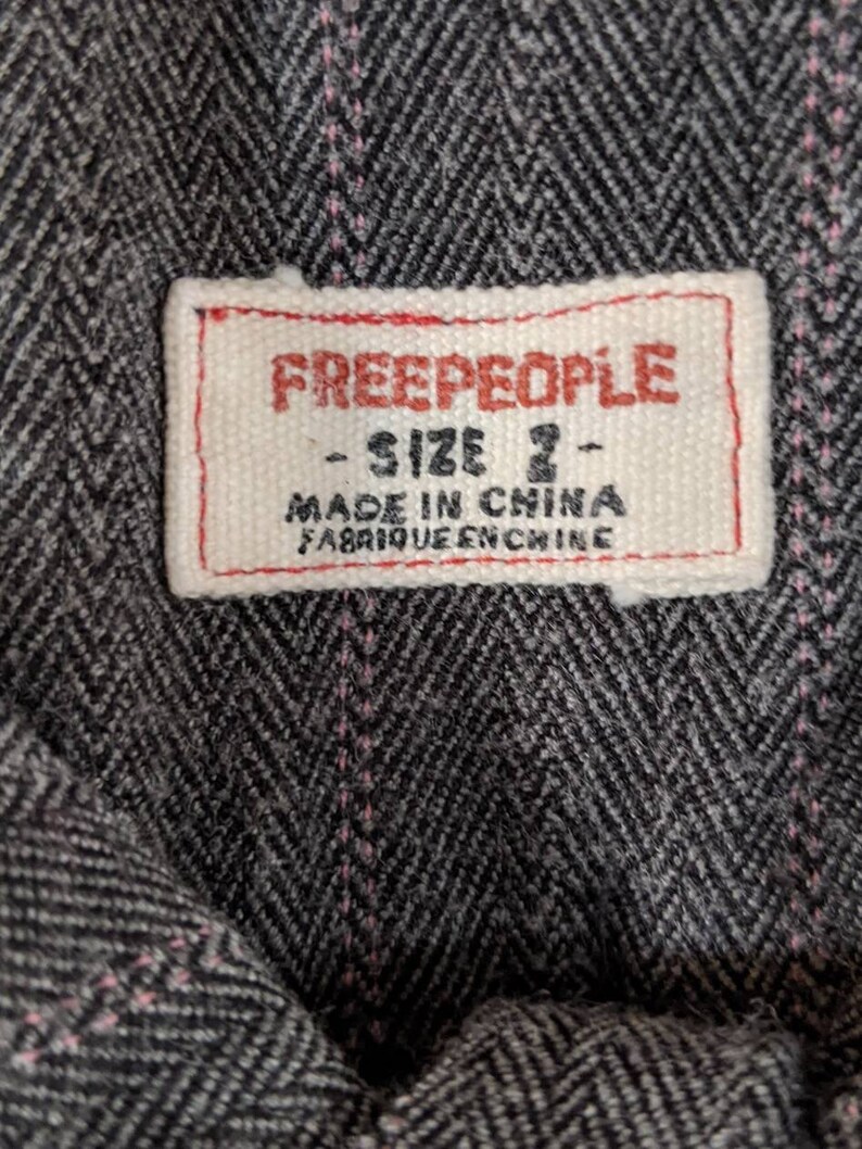 Peut inclure: Gros plan sur une &eacute;tiquette de tissu avec la marque "FREE PEOPLE" en lettres rouges. L'&eacute;tiquette indique &eacute;galement "SIZE 2" et "MADE IN CHINA FABRIQUE EN CHINE" en texte noir. Le tissu pr&eacute;sente un motif &agrave; chevrons gris et noir avec des coutures roses.