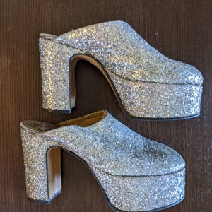 May include: A pair of silver glitter platform mules. These vintage-style shoes have a chunky heel and a thick platform sole. The open-back design and sparkling finish make them a statement piece. The heel height is approximately 4 inches.