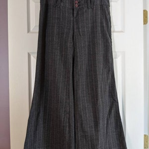 Peut inclure: Pantalon large &agrave; rayures grises fonc&eacute;es suspendu &agrave; une porte blanche. Le pantalon a une taille haute avec trois boutons marron et une fermeture &eacute;clair. Le tissu pr&eacute;sente des rayures verticales et une coupe &eacute;vas&eacute;e.