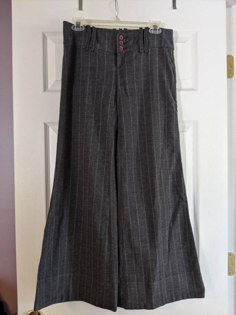 Peut inclure: Pantalon large &agrave; rayures grises fonc&eacute;es suspendu &agrave; une porte blanche. Le pantalon a une taille haute avec trois boutons marron et une fermeture &eacute;clair. Le tissu pr&eacute;sente des rayures verticales et une coupe &eacute;vas&eacute;e.