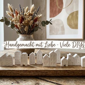 Könnte beinhalten: Eine Sammlung kleiner weißer Keramikhaus-Figuren, die auf einem Holztablett angeordnet sind. Die Häuser weisen verschiedene architektonische Details wie Fenster und Herzen auf. Der Text "Handgemacht mit Liebe • Viele DIYs" ist mittig eingeblendet.