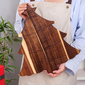 May include: A wooden cutting board in the shape of a Christmas tree. The board is made of dark brown wood with lighter wood accents. The tree-shaped board is held by a person wearing a blue shirt and apron.
