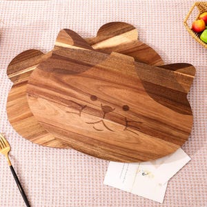 May include: Two wooden cutting boards shaped like a cat's face. The boards are made of light and dark brown wood with a carved cat face design. A gold fork and a small basket of fruit are in the background.