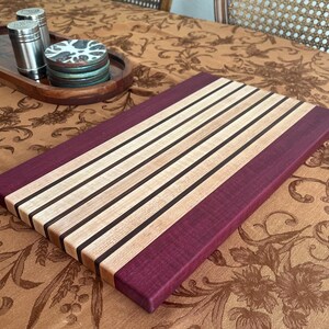 May include: A rectangular wooden cutting board with a striped design. The board features alternating light wood, dark wood, and purple wood stripes. The cutting board is approximately 18 inches long and 12 inches wide.