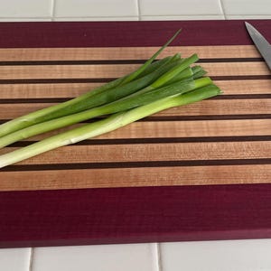 May include: A wooden cutting board with alternating stripes of light and dark wood, and a deep red border. Green onions and a silver knife rest on the board, ready for food preparation. Kitchen and cooking supplies.