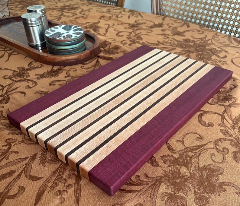 May include: A rectangular wooden cutting board with a striped design. The board features alternating light wood, dark wood, and purple wood stripes. The cutting board is approximately 18 inches long and 12 inches wide.