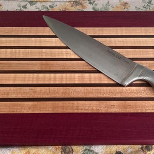 May include: A rectangular wooden cutting board with alternating stripes of light and dark wood, and a deep red border. A stainless steel chef's knife rests diagonally across the board, ready for use. Kitchen and dining.