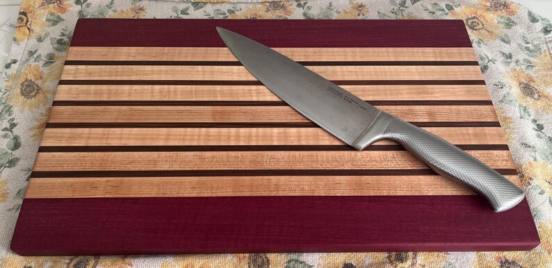 May include: A rectangular wooden cutting board with alternating stripes of light and dark wood, and a deep red border. A stainless steel chef's knife rests diagonally across the board, ready for use. Kitchen and dining.