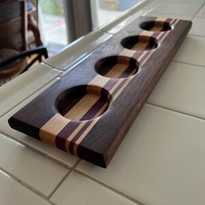 May include: A rectangular wooden beer flight paddle featuring four circular cutouts for glasses. The board is constructed from alternating strips of dark brown, light tan, and deep purple wood, creating a striped pattern across the surface.