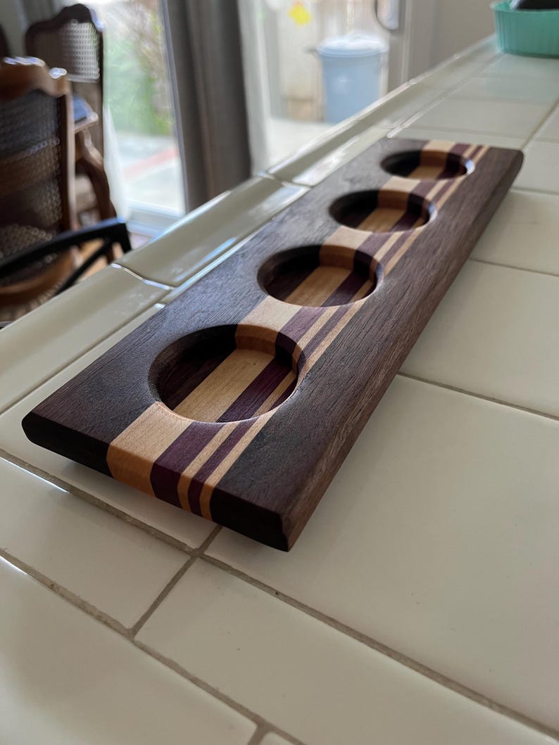 May include: A rectangular wooden beer flight paddle featuring four circular cutouts for glasses. The board is constructed from alternating strips of dark brown, light tan, and deep purple wood, creating a striped pattern across the surface.