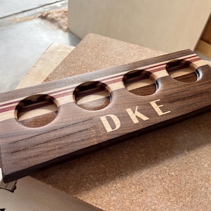 May include: A rectangular wooden beer flight paddle featuring three circular cutouts for glasses. The board is crafted from dark walnut wood with horizontal stripes of lighter wood and purple accents. The letters DKE are engraved on the front.