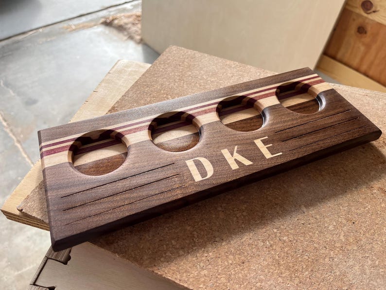 May include: A rectangular wooden beer flight paddle featuring three circular cutouts for glasses. The board is crafted from dark walnut wood with horizontal stripes of lighter wood and purple accents. The letters DKE are engraved on the front.
