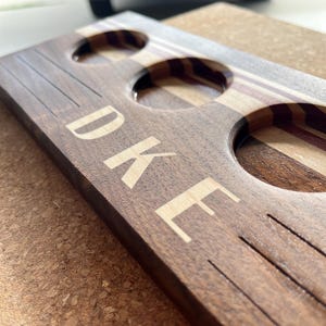 May include: A rectangular dark wood serving board featuring three circular cutouts for glasses. The board displays a light-colored wood inlay stripe and the engraved text "DUKE" in a clean, sans-serif font, accompanied by etched horizontal lines.