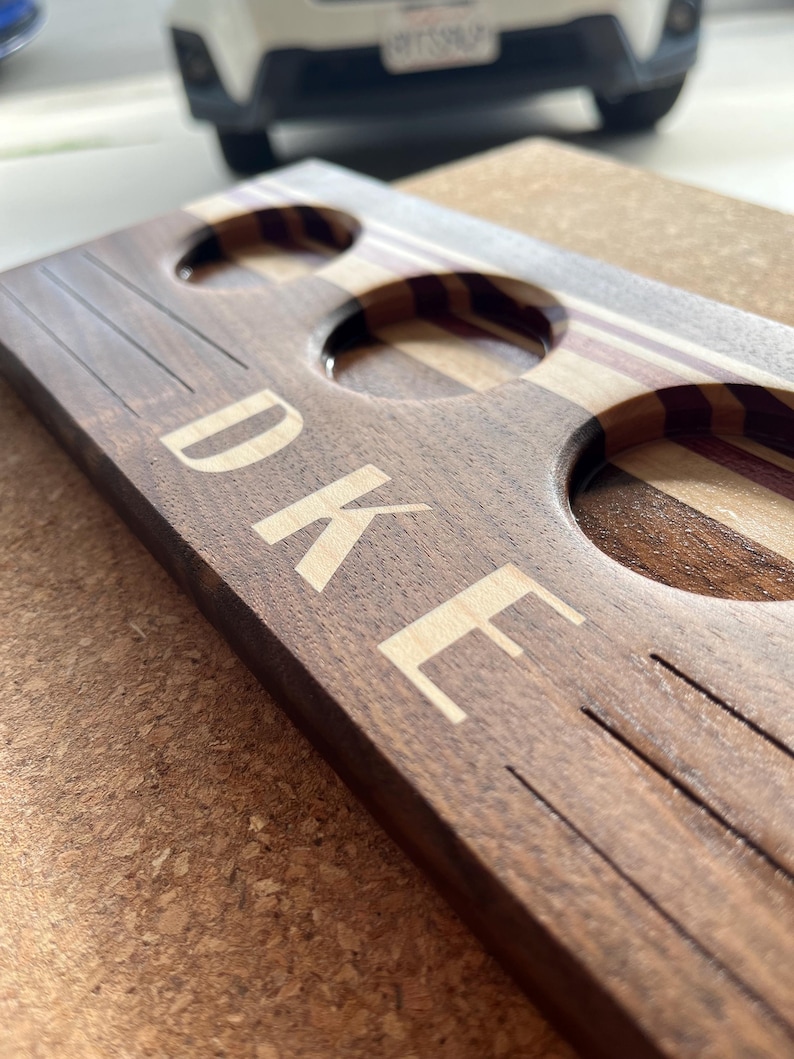 May include: A rectangular dark wood serving board featuring three circular cutouts for glasses. The board displays a light-colored wood inlay stripe and the engraved text "DUKE" in a clean, sans-serif font, accompanied by etched horizontal lines.