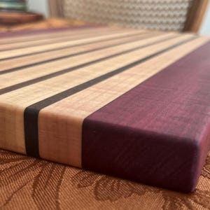 May include: A handcrafted wooden cutting board with alternating light and dark wood stripes, and a solid purple-hued end. The board is rectangular and appears to be made of high-quality wood, suitable for kitchen use.