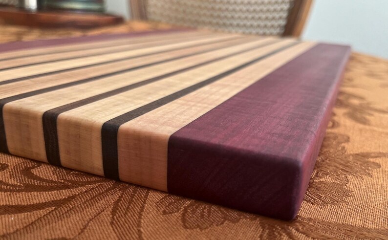 May include: A handcrafted wooden cutting board with alternating light and dark wood stripes, and a solid purple-hued end. The board is rectangular and appears to be made of high-quality wood, suitable for kitchen use.
