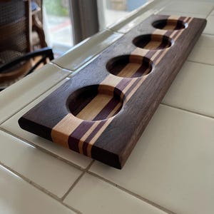 May include: A rectangular wooden serving paddle featuring four circular cutouts for glasses. The board is constructed from alternating strips of dark brown, light tan, and deep purple wood, creating a striped pattern across the surface.