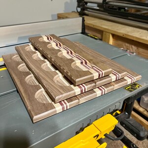 May include: A stack of three rectangular wooden serving boards, each featuring four circular indentations. The boards are crafted from dark brown wood with contrasting light tan and deep purple striped inlay accents running along the length.