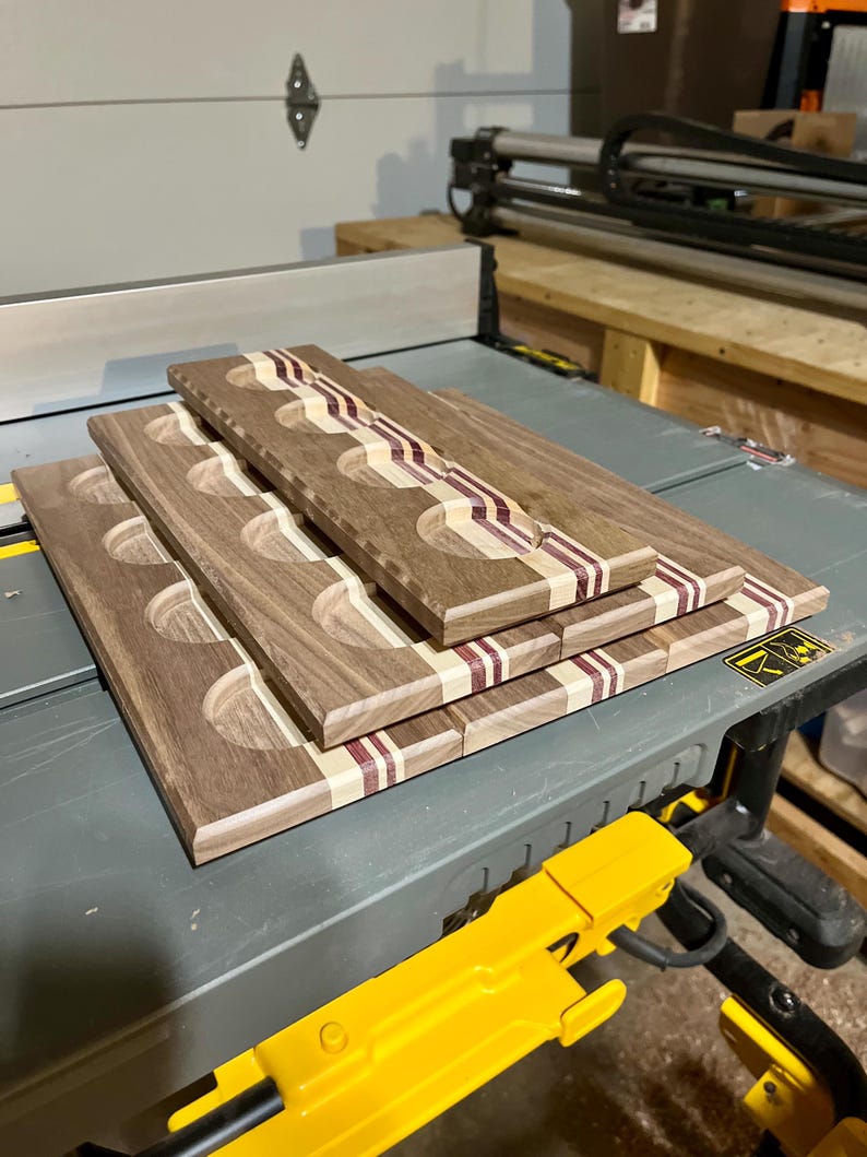 May include: A stack of three rectangular wooden serving boards, each featuring four circular indentations. The boards are crafted from dark brown wood with contrasting light tan and deep purple striped inlay accents running along the length.