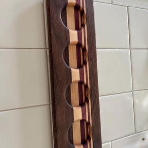May include: A rectangular wooden beer flight paddle featuring four circular cutouts for glasses. The board is constructed from alternating vertical strips of dark brown walnut, light maple, and deep purple heart wood, creating a striped pattern.