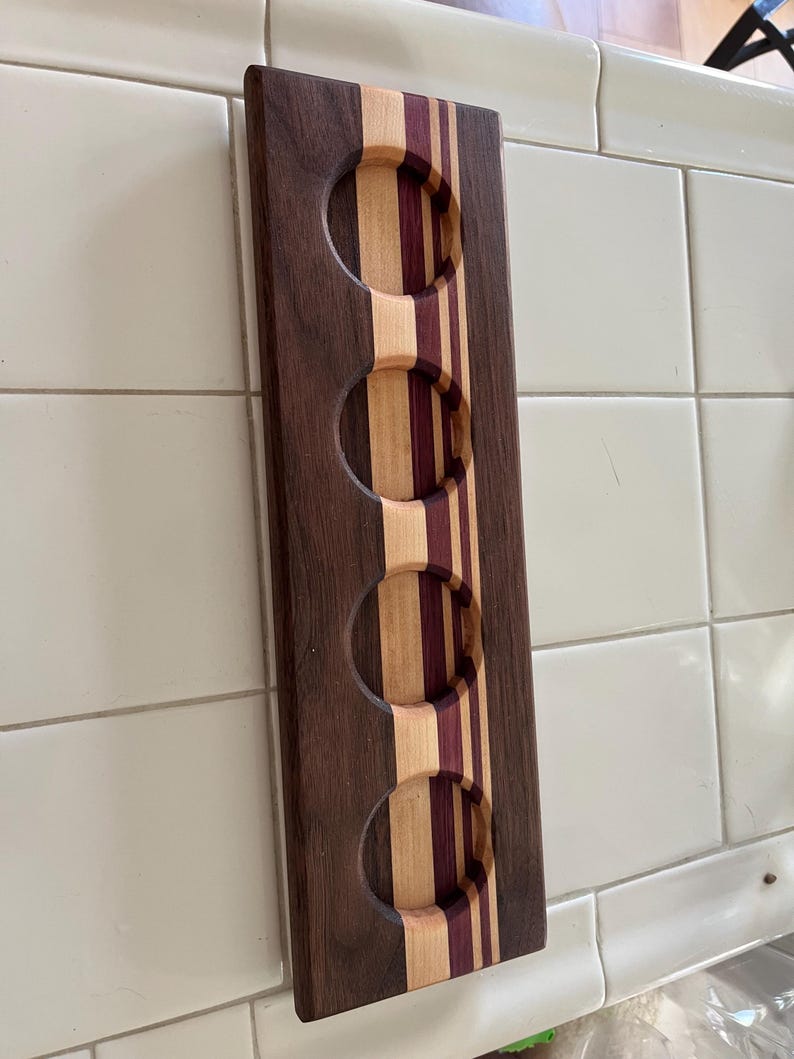 May include: A rectangular wooden beer flight paddle featuring four circular cutouts for glasses. The board is constructed from alternating vertical strips of dark brown walnut, light maple, and deep purple heart wood, creating a striped pattern.