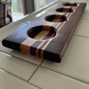 May include: A rectangular wooden serving paddle featuring four circular cutouts for glasses. The board is constructed from alternating strips of dark brown, light tan, and deep purple wood, showcasing a smooth, polished finish throughout the piece.