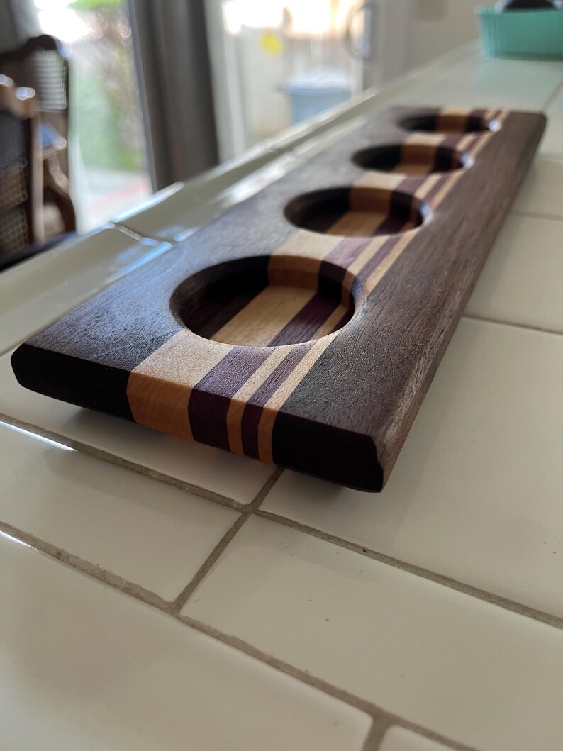 May include: A rectangular wooden serving paddle featuring four circular cutouts for glasses. The board is constructed from alternating strips of dark brown, light tan, and deep purple wood, showcasing a smooth, polished finish throughout the piece.