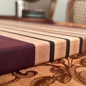 May include: A rectangular wooden cutting board with alternating light and dark wood stripes. The end of the board is a deep purple color. The board has a smooth, finished surface and is likely used for food preparation.