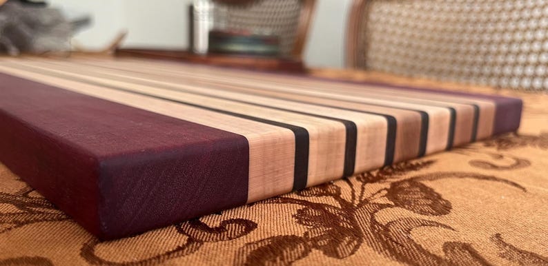 May include: A rectangular wooden cutting board with alternating light and dark wood stripes. The end of the board is a deep purple color. The board has a smooth, finished surface and is likely used for food preparation.
