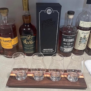 May include: A wooden flight board with four clear glass tasting glasses arranged in a row. Behind the board are five bottles of whiskey, including labels for Frey Ranch, Weller Special Reserve, Union Horse 10 Year, Rebel 10 Year, and Willett.
