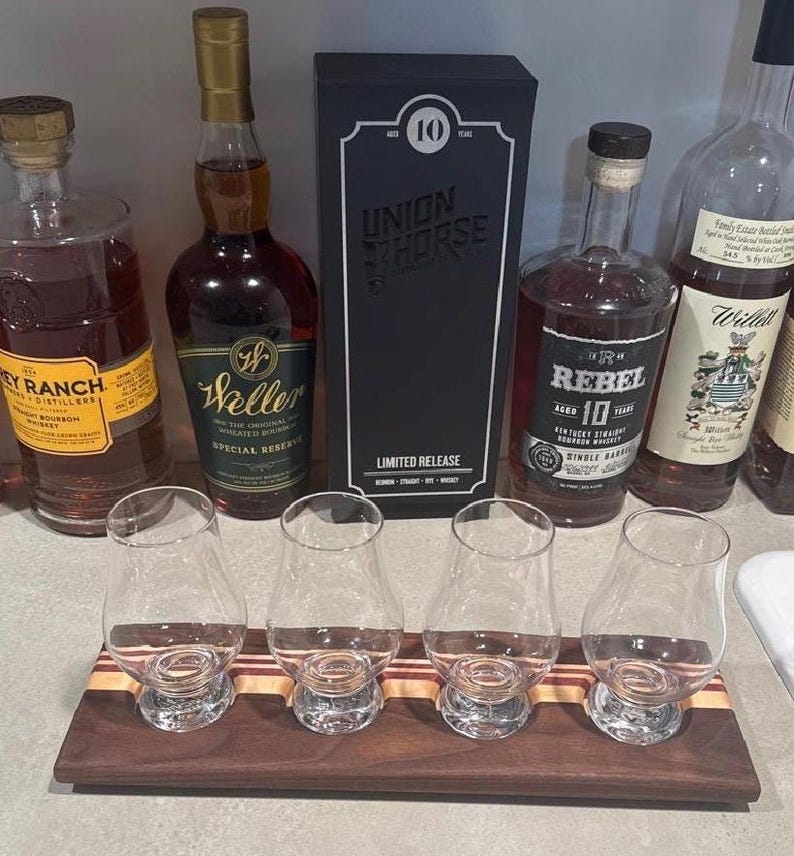 May include: A wooden flight board with four clear glass tasting glasses arranged in a row. Behind the board are five bottles of whiskey, including labels for Frey Ranch, Weller Special Reserve, Union Horse 10 Year, Rebel 10 Year, and Willett.