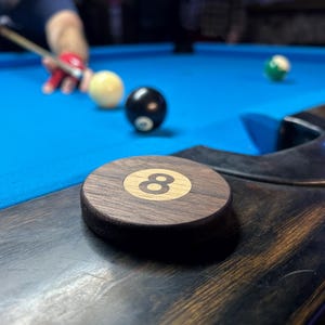 May include: A circular wooden marker featuring a light-colored center inlay with the number eight in black, resting on the dark wood rail of a blue pool table. In the background, a pool cue is positioned near a white cue ball and an eight ball.