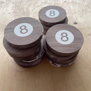 May include: Three stacks of circular wooden coasters, each featuring a white circular inlay with the number eight printed in brown. The coasters are made of dark brown wood with a visible grain, and the stacks are arranged in a triangular formation.