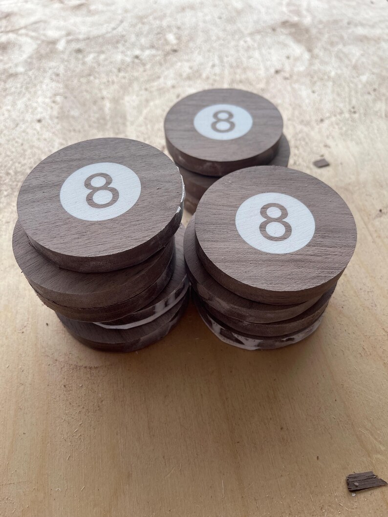 May include: Three stacks of circular wooden coasters, each featuring a white circular inlay with the number eight printed in brown. The coasters are made of dark brown wood with a visible grain, and the stacks are arranged in a triangular formation.