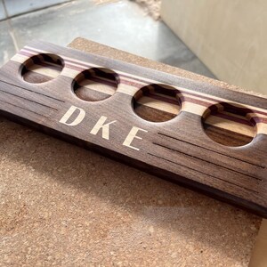 May include: A rectangular wooden beer flight paddle featuring four circular cutouts for glasses. The board is crafted from dark brown wood with light and purple-toned wood accents. The letters DKE are engraved in the center with horizontal lines.